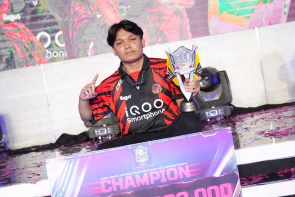 Seorang pemain e-sports mengenakan jersey hitam merah memegang trofi juara sambil duduk di panggung dengan latar tulisan CHAMPION.