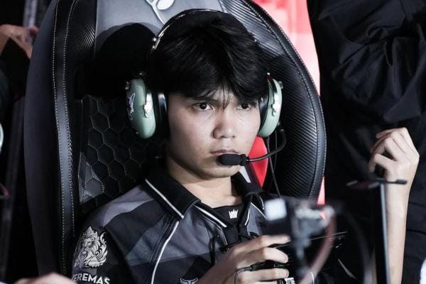 Seorang pemain e-sports mengenakan headset dan seragam tim hitam abu-abu sedang fokus di depan layar saat pertandingan berlangsung.