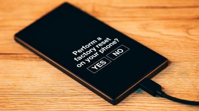 Layar ponsel menampilkan pesan konfirmasi untuk melakukan factory reset dengan pilihan tombol Yes dan No di atas meja kayu.