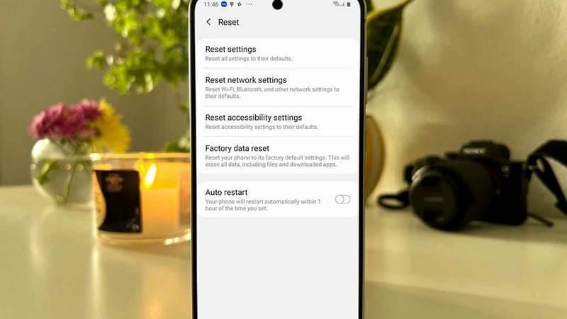 Layar ponsel menampilkan menu pengaturan reset termasuk factory data reset, dengan latar belakang meja berisi lilin dan kamera.
