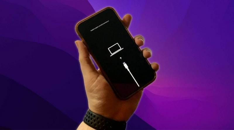 Seseorang memegang iPhone yang menampilkan layar mode pemulihan dengan ikon kabel dan komputer di latar belakang ungu.