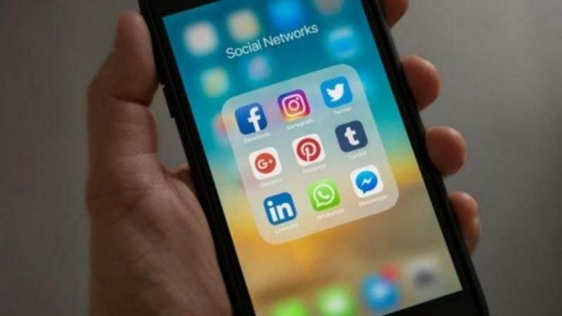 Seseorang memegang ponsel pintar yang menampilkan folder aplikasi media sosial seperti Facebook, Instagram, Twitter, dan WhatsApp di layar.