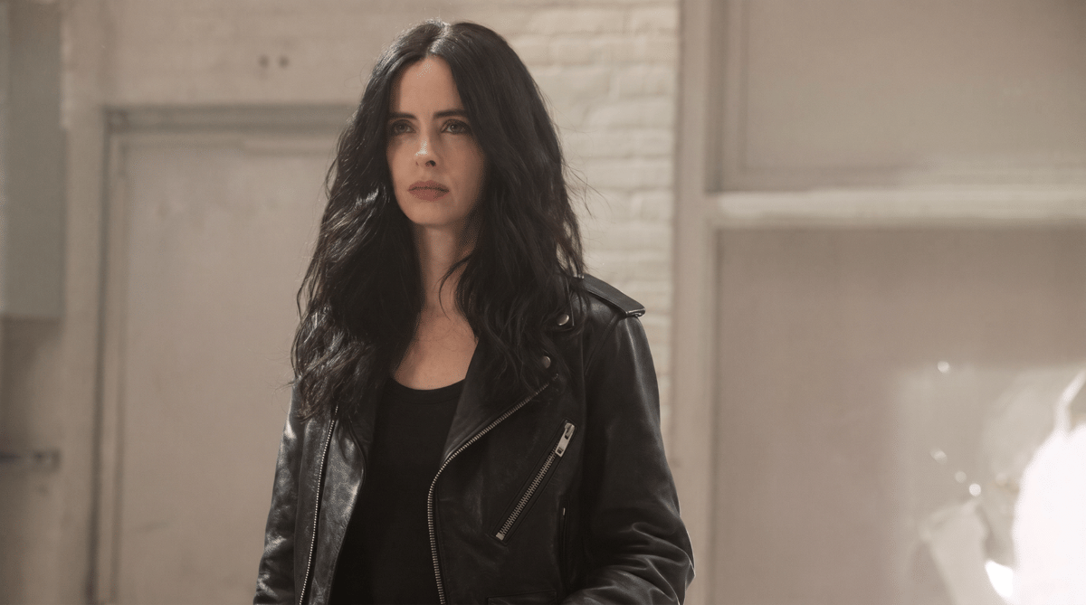 Seorang wanita berambut hitam panjang mengenakan jaket kulit hitam berdiri di dalam ruangan dengan dinding bata terang.