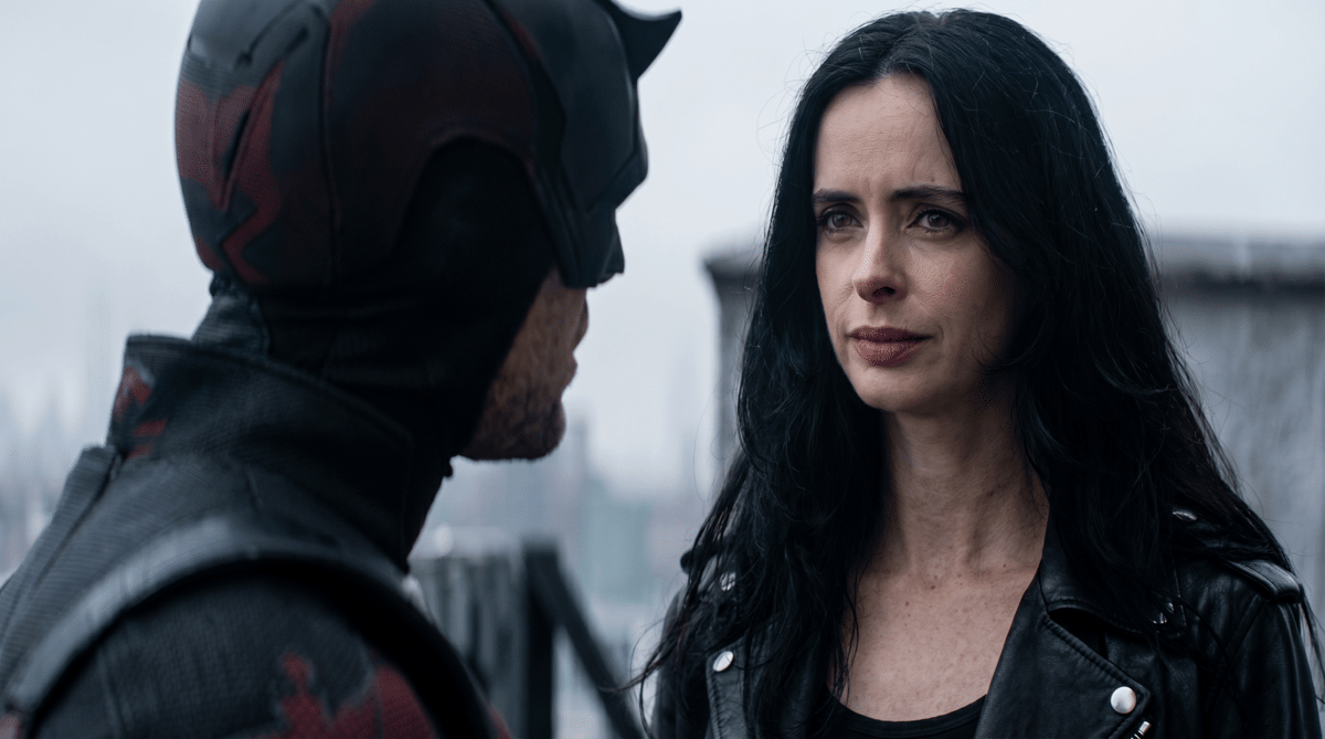 Daredevil mengenakan kostum merah hitam berbicara dengan Jessica Jones berjaket kulit hitam di latar kota berkabut.