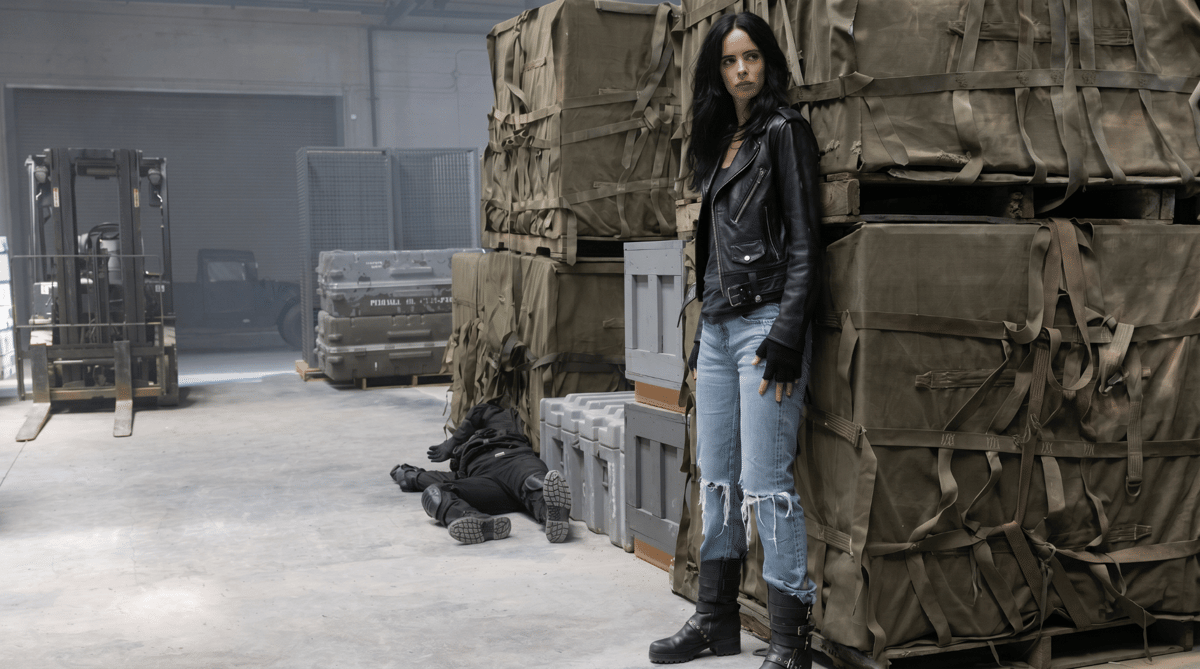 Seorang wanita berjaket kulit hitam dan celana jeans robek berdiri di gudang dengan tumpukan peti besar dan seorang pria tergeletak di lantai.