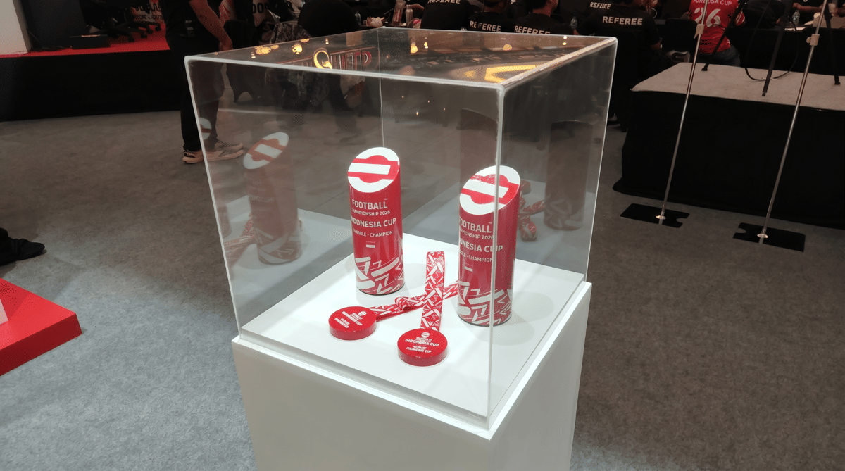 Medali dan piala eFootball Indonesia Cup 2026 berwarna merah putih dipajang dalam kotak kaca di atas podium putih di area acara.